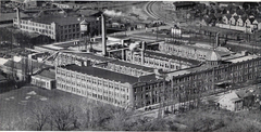 Ames Street Plant & Campus (Aerial View) Comprehensive view of the 300,000+ sq. ft. complex showing the rooftop weather observatory dome, water tower, rail spur, and employee athletic field in the foreground.