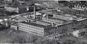 Ames Street Plant & Campus (Aerial Overview) Comprehensive view showing the multi-story comb-tooth wings, rooftop weather dome, water tower, and foreground athletic field.