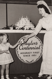Centennial Cake & Refreshments Hostess in 1851 gown serving ice cream to a young guest in front of the multi-tiered 100th anniversary cake.